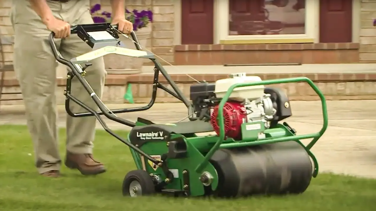 Person using a green lawn aerator machine on grass in front of a house with brick steps.