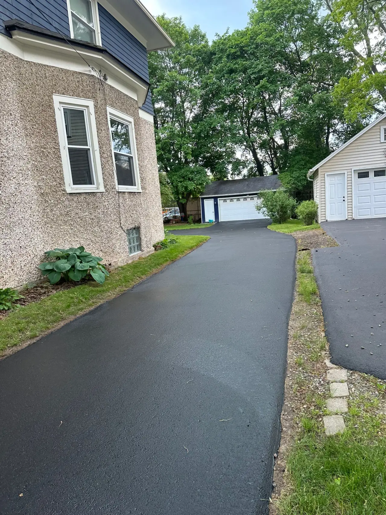 Freshly paved driveway by a house, leading to a detached garage, with trees in the background.