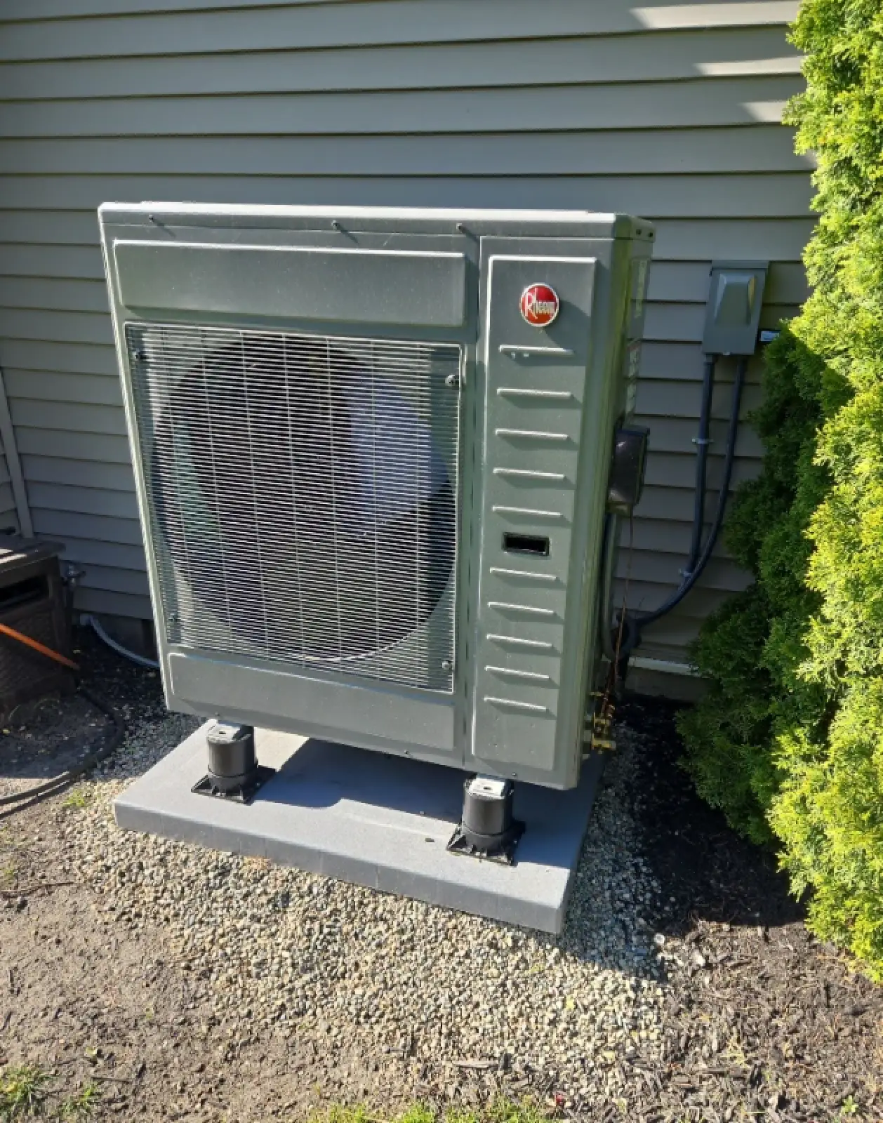 A Rheem heat pump unit sits on a pad next to a house with gray siding and a green shrub.