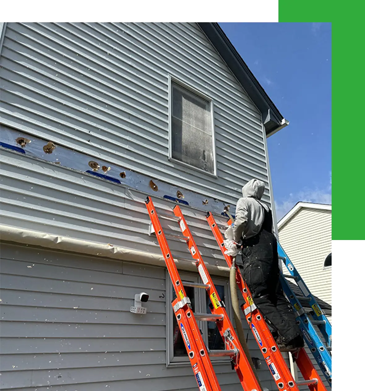 Person on ladders repairing damaged siding on a two-story house under a partly cloudy sky.