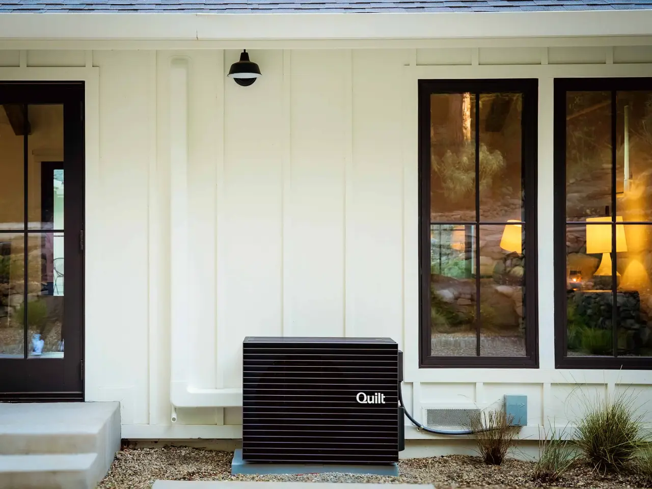 A Quilt heat pump is installed outside a modern white house near two large windows.
