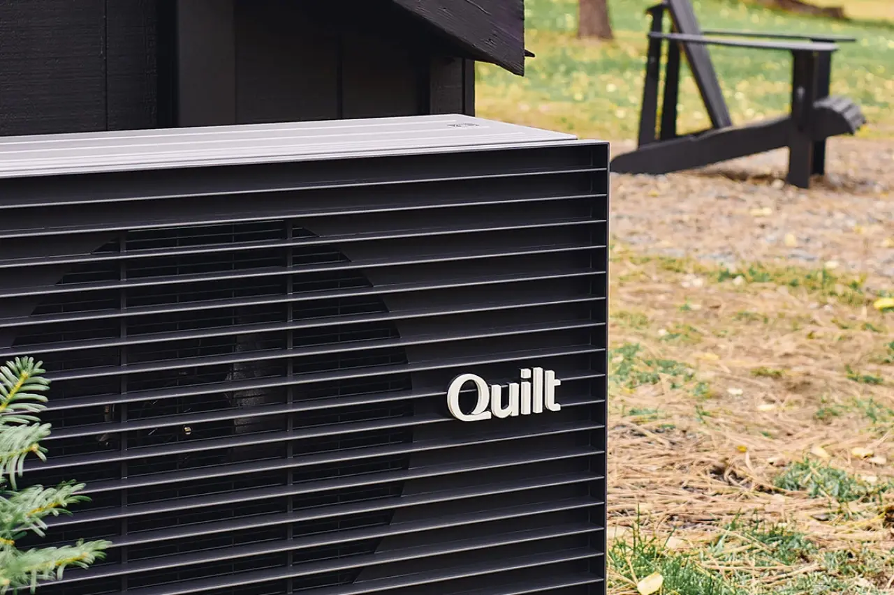 A black outdoor heat pump unit labeled Quilt sits next to a building with grass nearby.