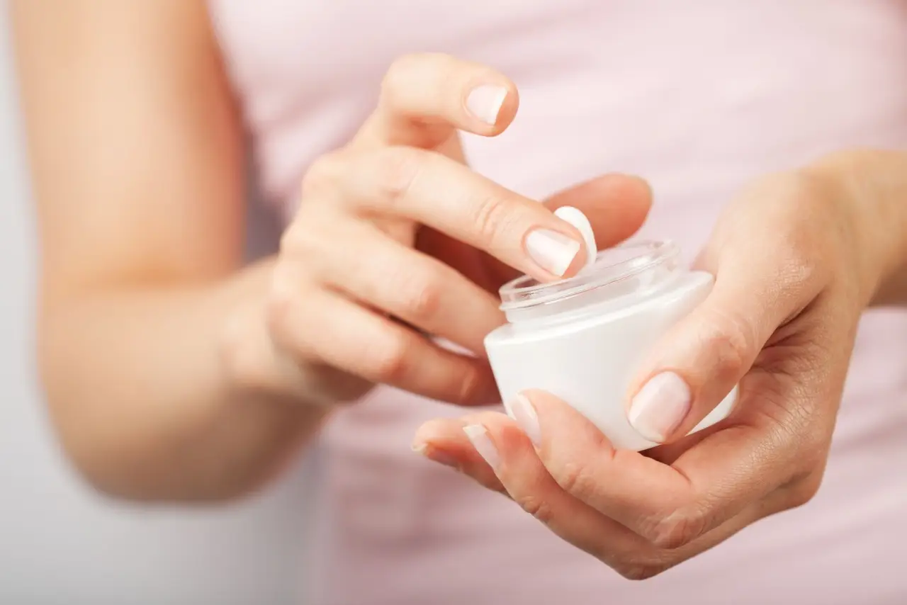 Hands scooping moisturizer from a small white jar.