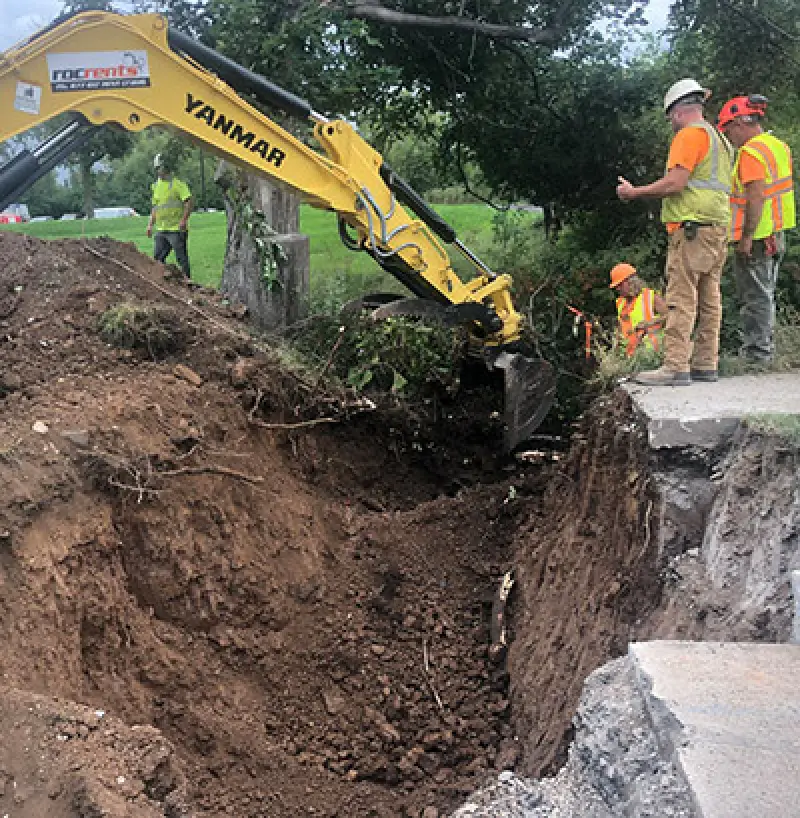 A yellow excavator digs a deep hole while construction workers in safety gear stand nearby.