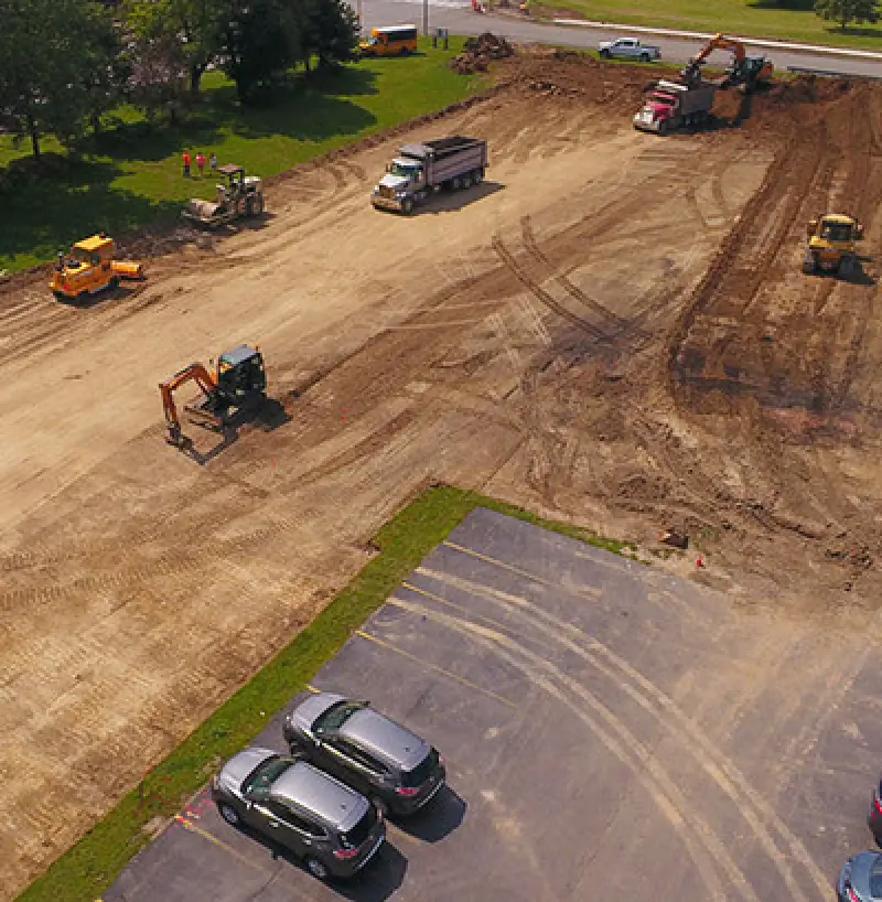 Aerial view of a construction site with vehicles, equipment, and a nearby parking lot.