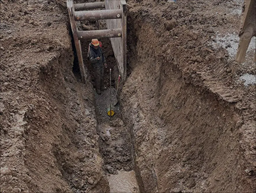 A worker with a hard hat stands in a deep, muddy trench supported by wooden beams.