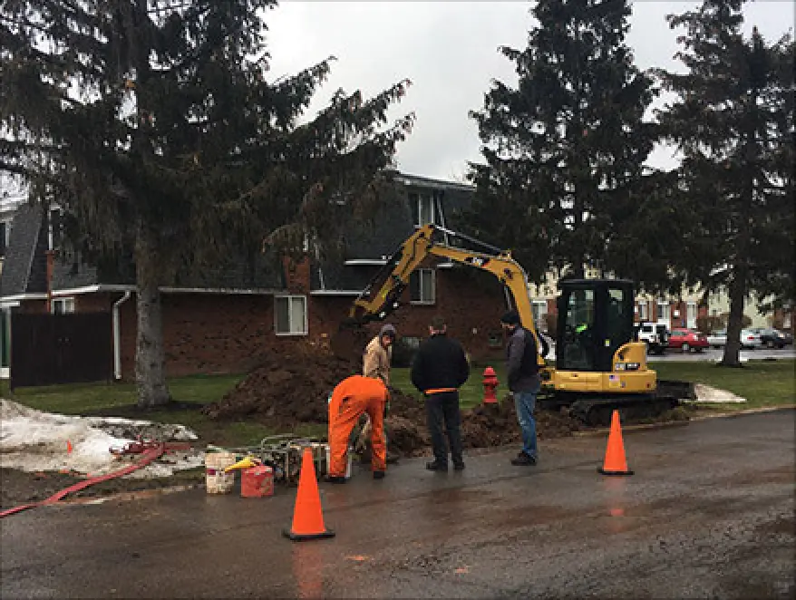 Four workers with an excavator dig near a house beside traffic cones on a cloudy day.