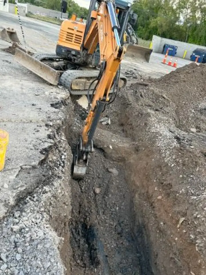 Excavator digging a deep trench in a construction site with gravel and dirt piles nearby.