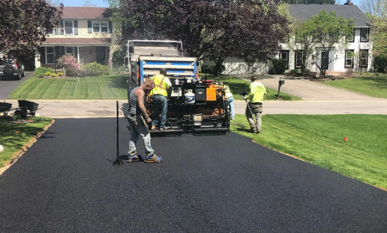 Workers paving a residential driveway with asphalt; houses and green lawns in the background.