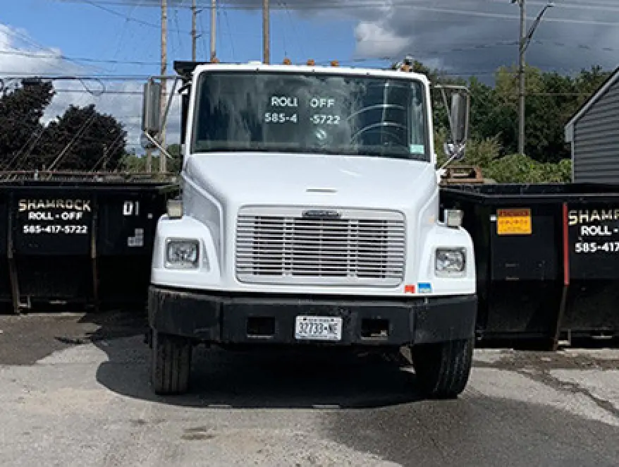 White roll-off truck parked in front of two black Shamrock dumpsters on a cloudy day.
