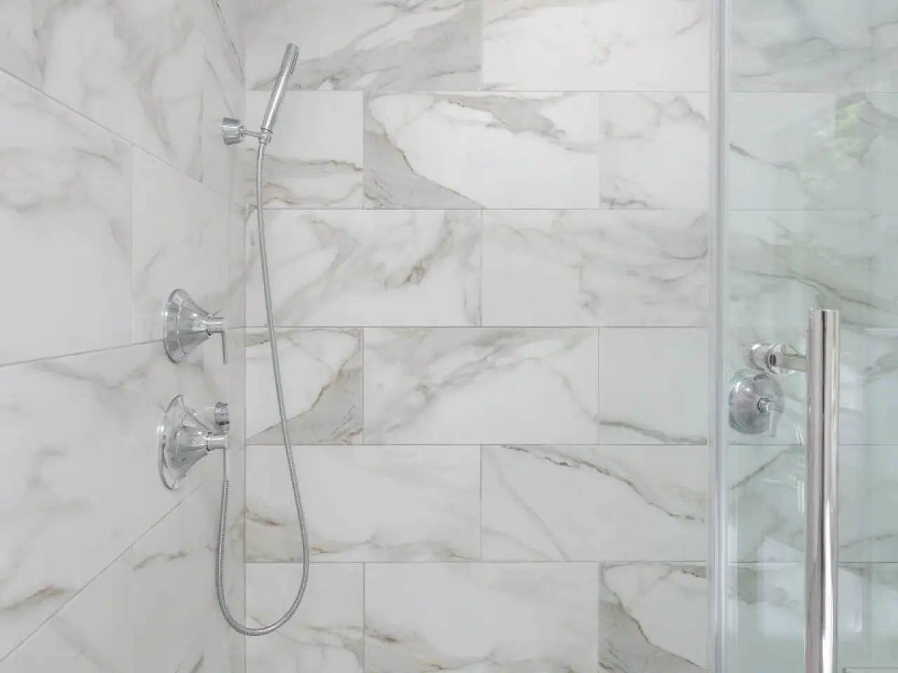 Modern shower with marble tile walls, chrome fixtures, handheld showerhead, and glass door.