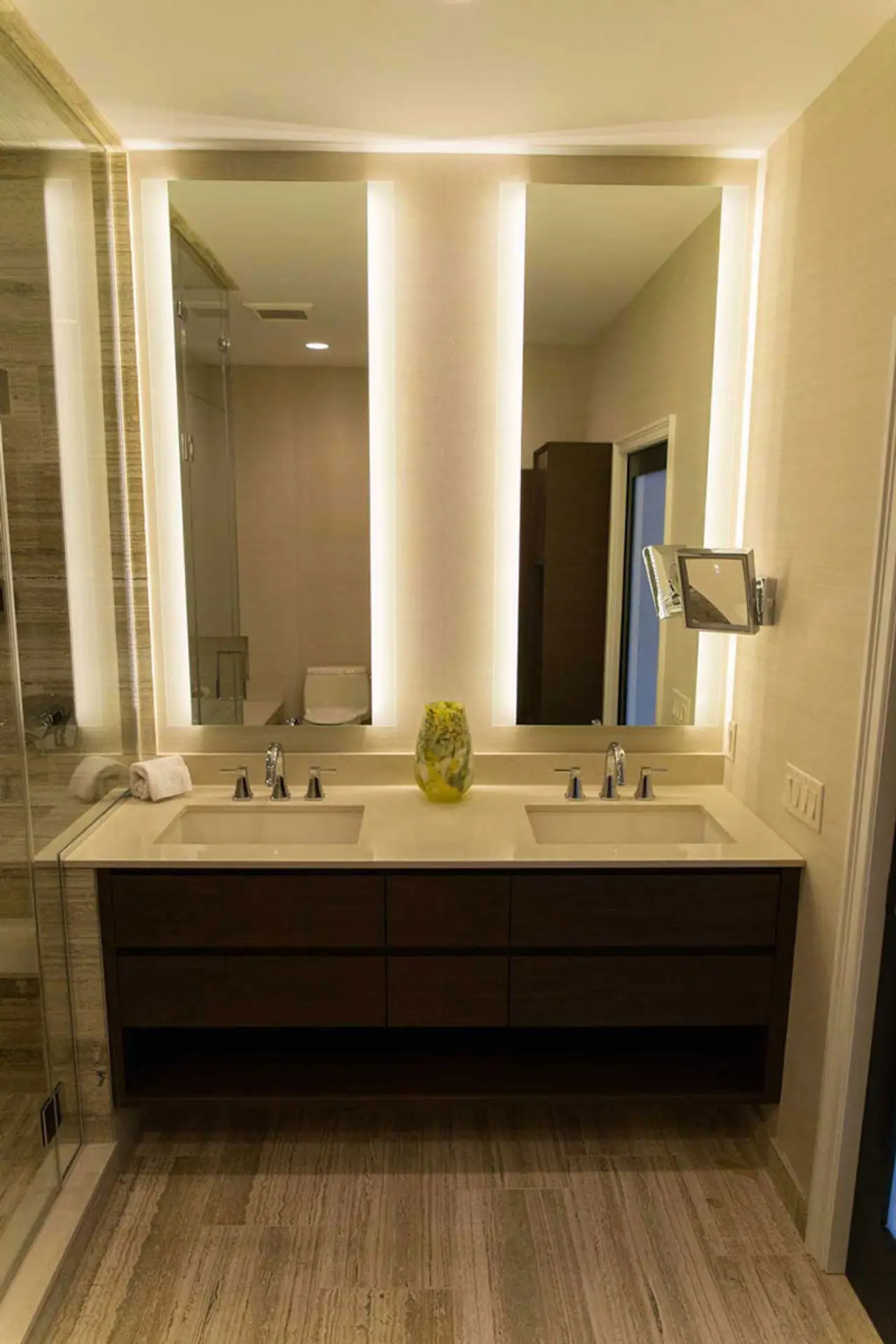 Dark wood double vanity with illuminated mirrors beside a glass-enclosed travertine shower.