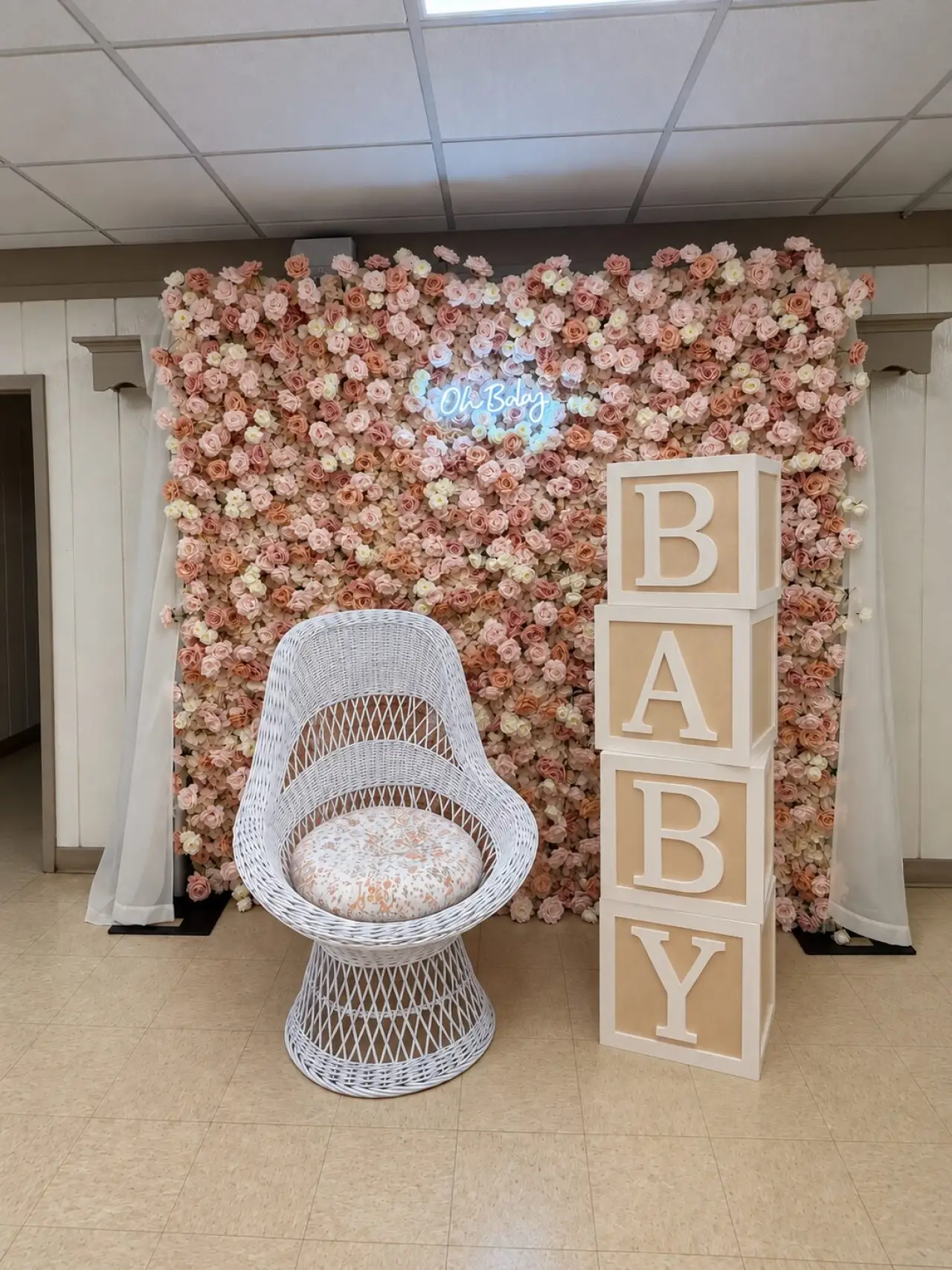 Stacked BABY block letters beside a white wicker chair in front of a blush flower wall with an Oh Baby neon sign
