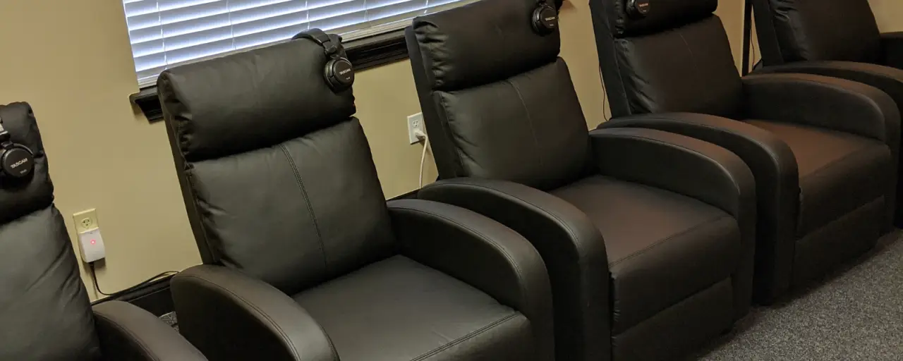 Four black leather chairs with headphones rest against their headrests, next to window blinds.