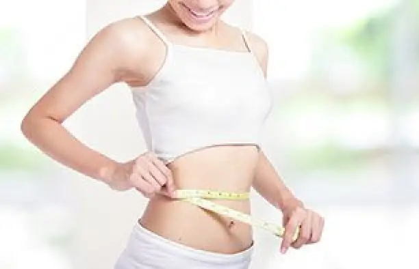 Woman in white clothing measuring her waist with a tape measure, smiling, indoors.