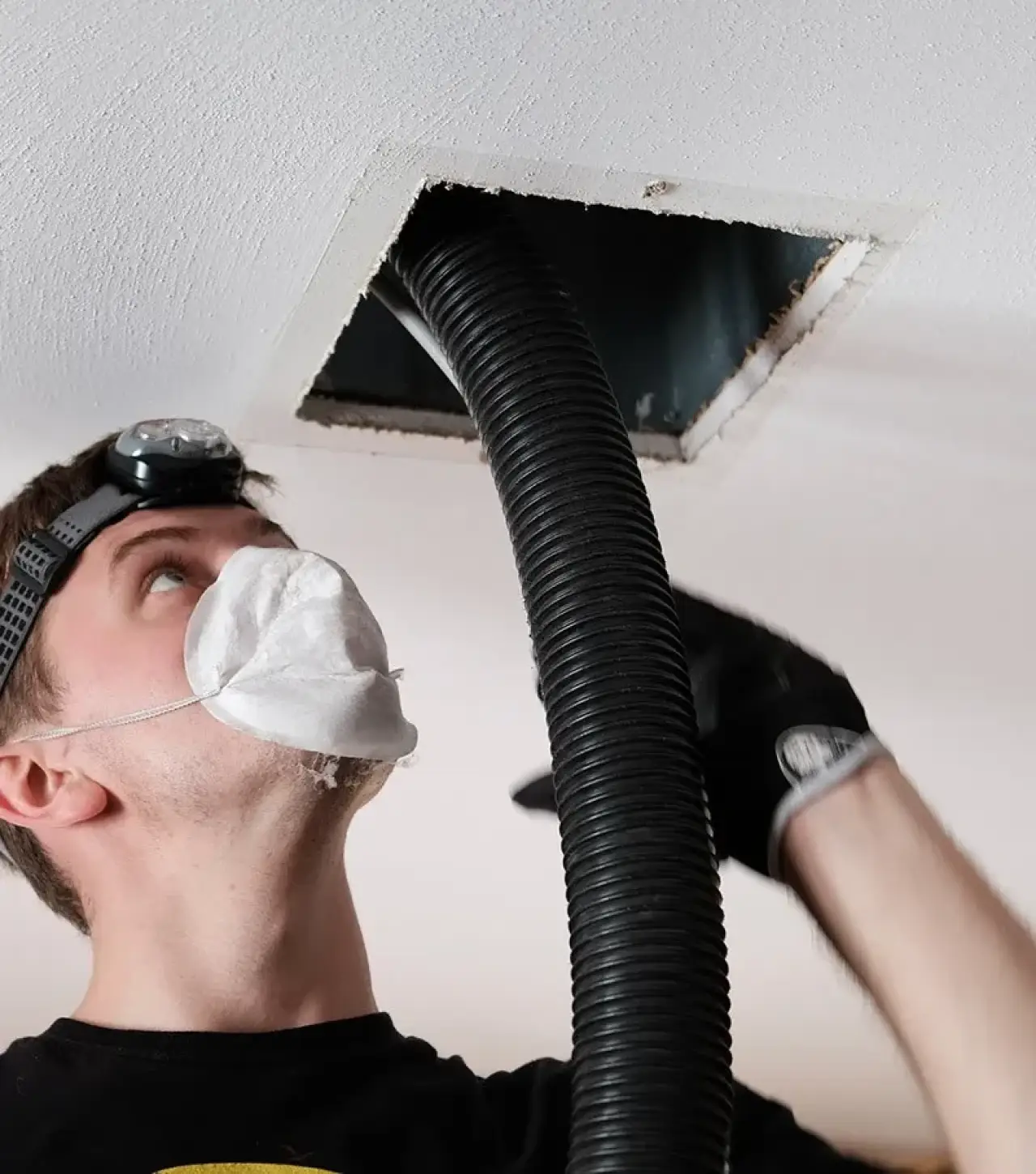 A man wearing a mask and headlamp using a tube to clear out a vent in the ceiling