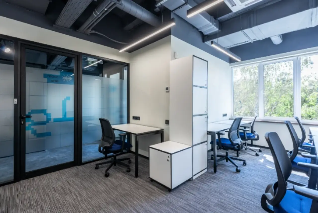 Modern office with desks, blue chairs, large windows, and a glass-walled meeting room.