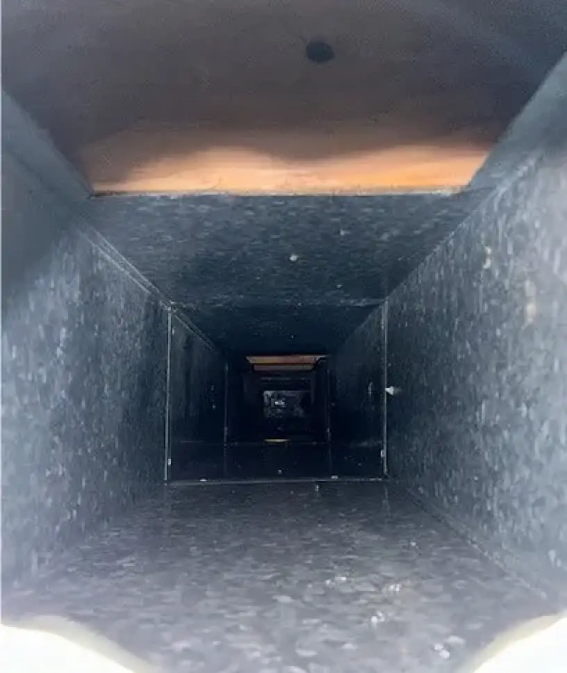 View looking down the inside of a rectangular air duct with dim lighting and dust.