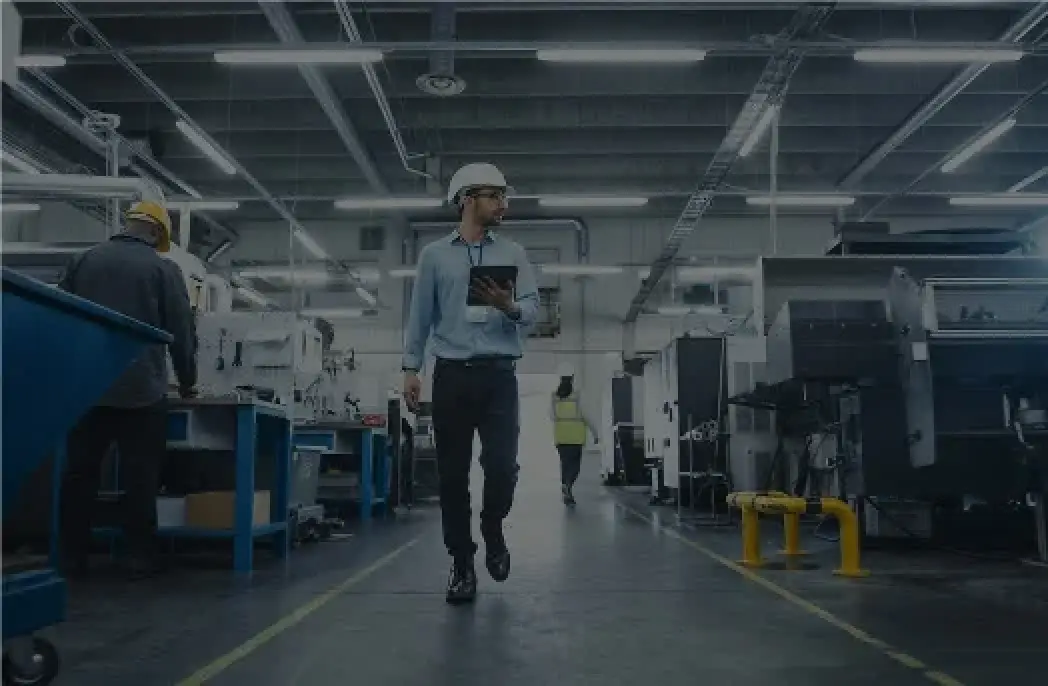 Man in hard hat walks through factory holding tablet; other workers and machinery are visible.