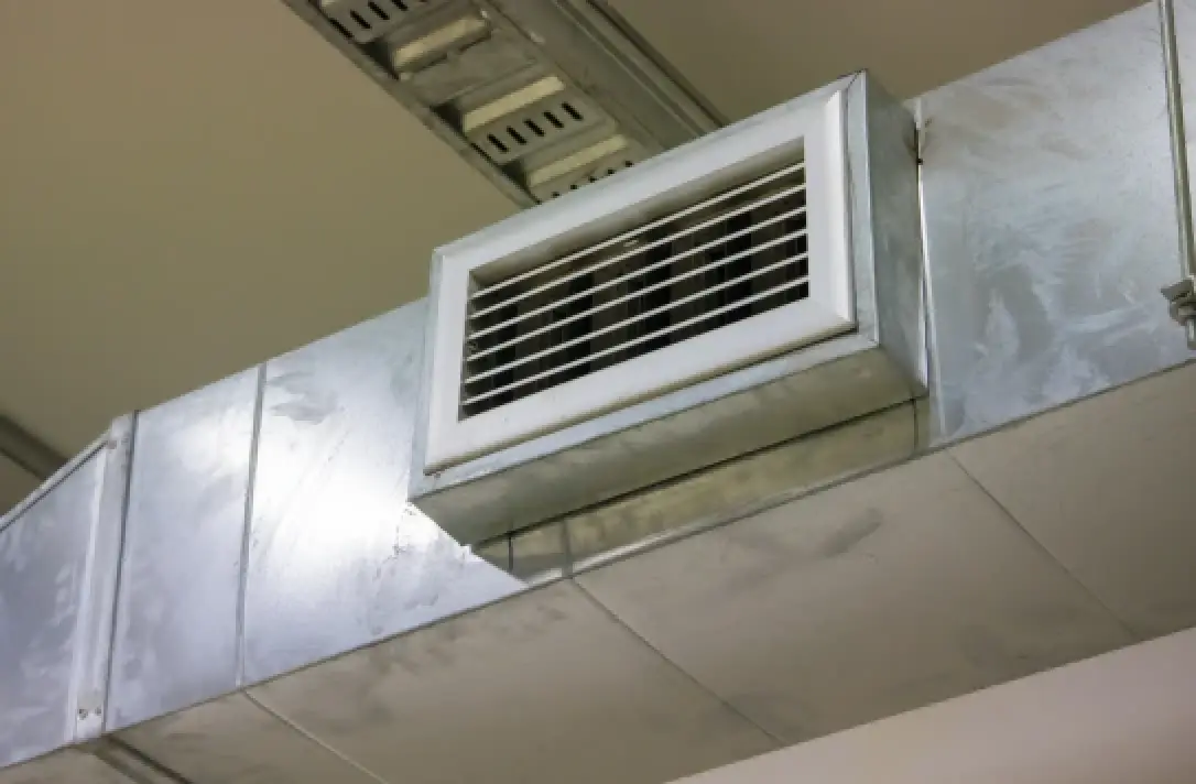 A metal air vent with a white grille on a ceiling-mounted duct in an indoor space.