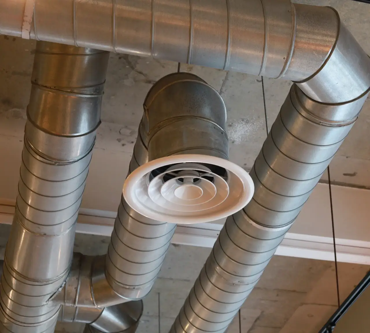 Exposed metal air ducts and a circular air vent on a ceiling with concrete and panels.