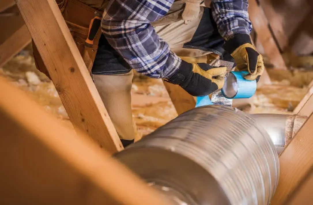 Person in gloves insulating an air duct in an attic with wooden beams and insulation.
