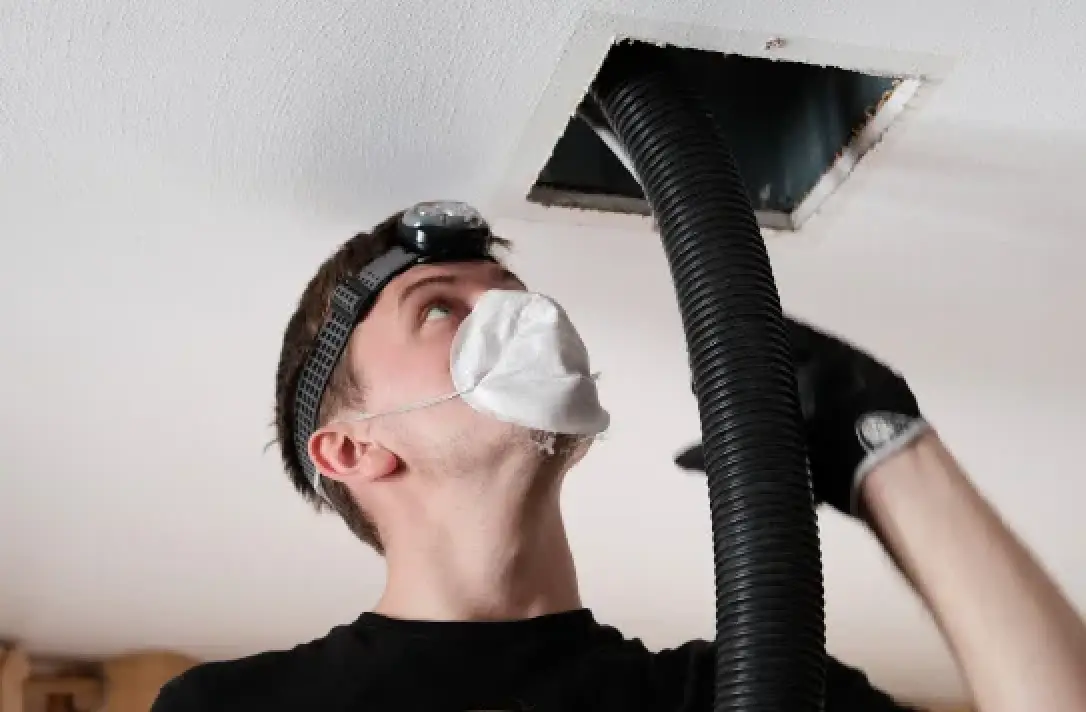Man wearing a mask and headlamp cleaning an air duct with a large vacuum hose.