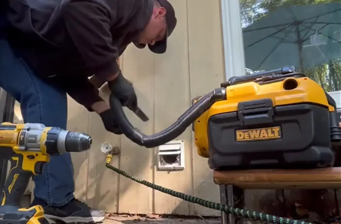 Person using a DeWalt vacuum to clean a dryer vent next to a yellow drill outdoors.
