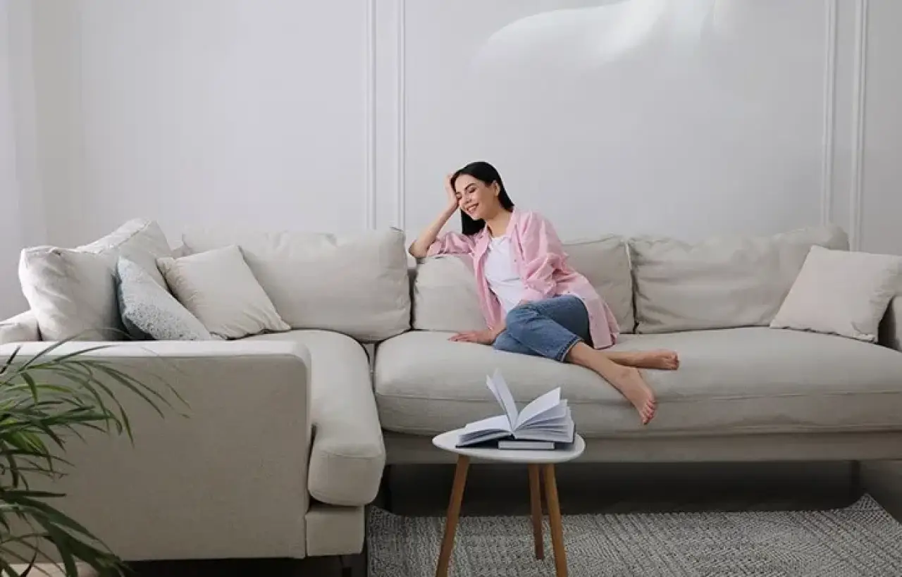Woman relaxing on a beige sofa, smiling, with an open book on a small round table nearby.