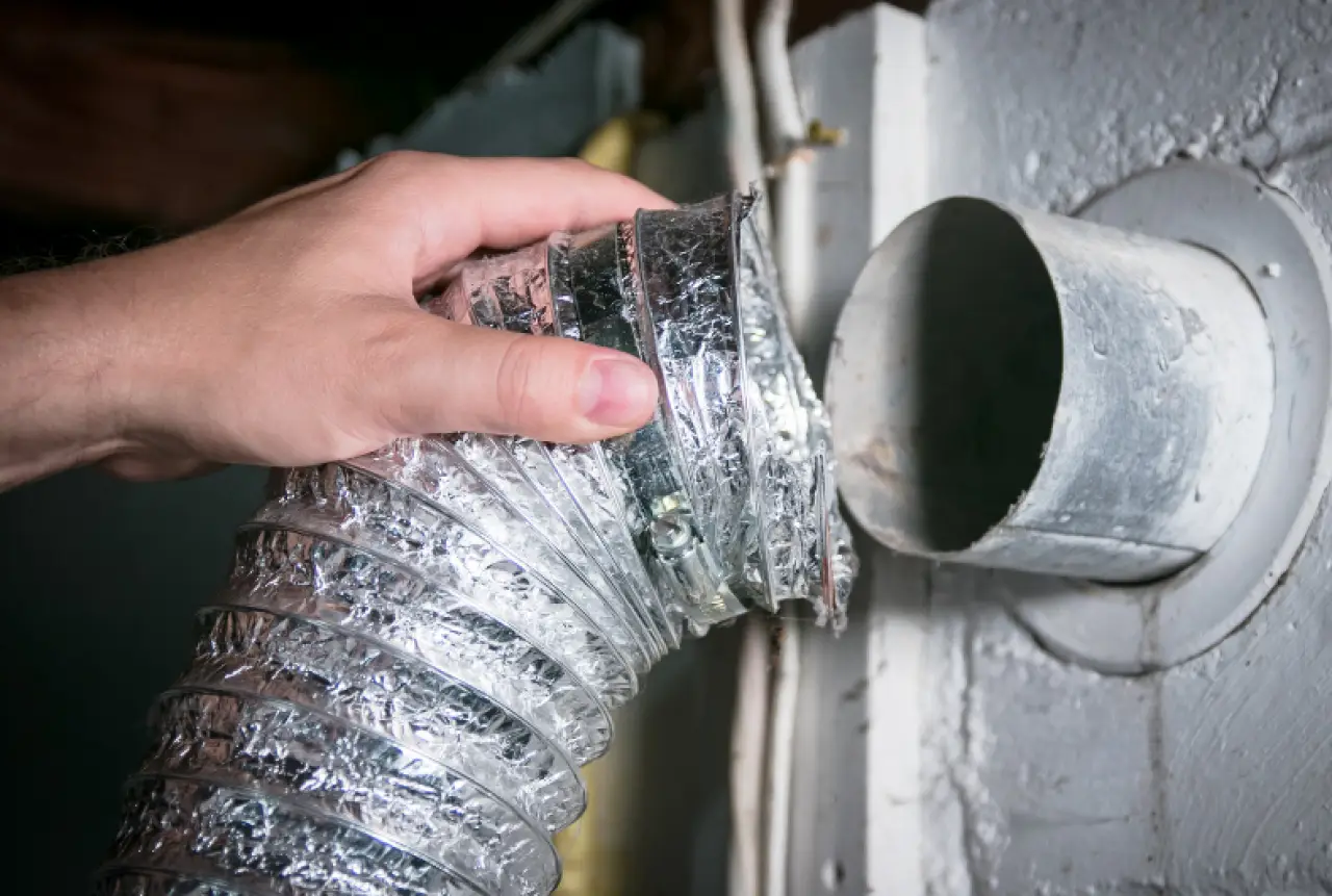 A hand connects a flexible dryer vent hose to a round metal exhaust duct on a wall.