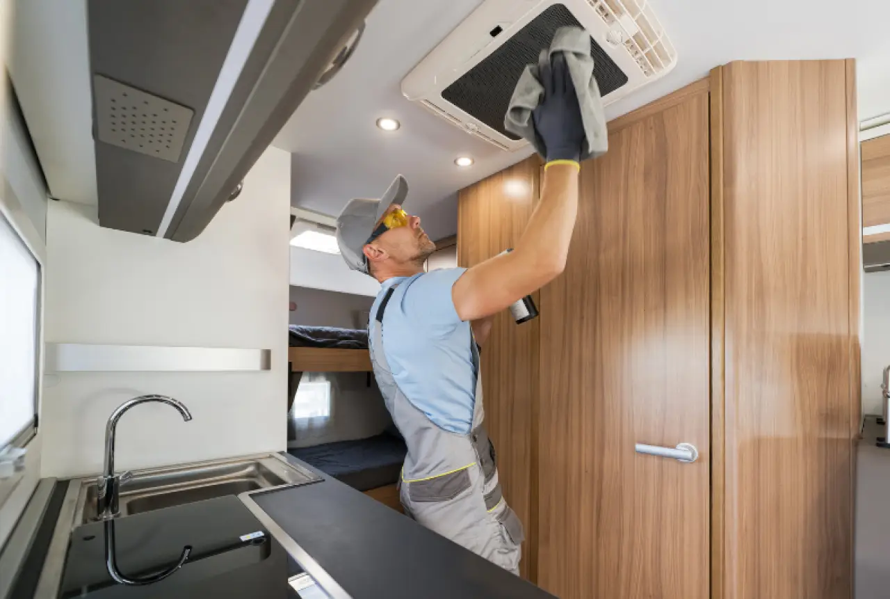 A man cleans an air conditioning vent inside a modern RV with wooden cabinets.