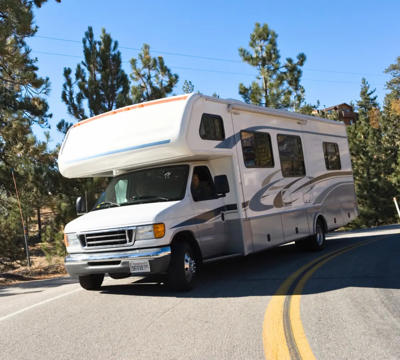 A white RV driving around a sharp curve on a forested road with tall pine trees.