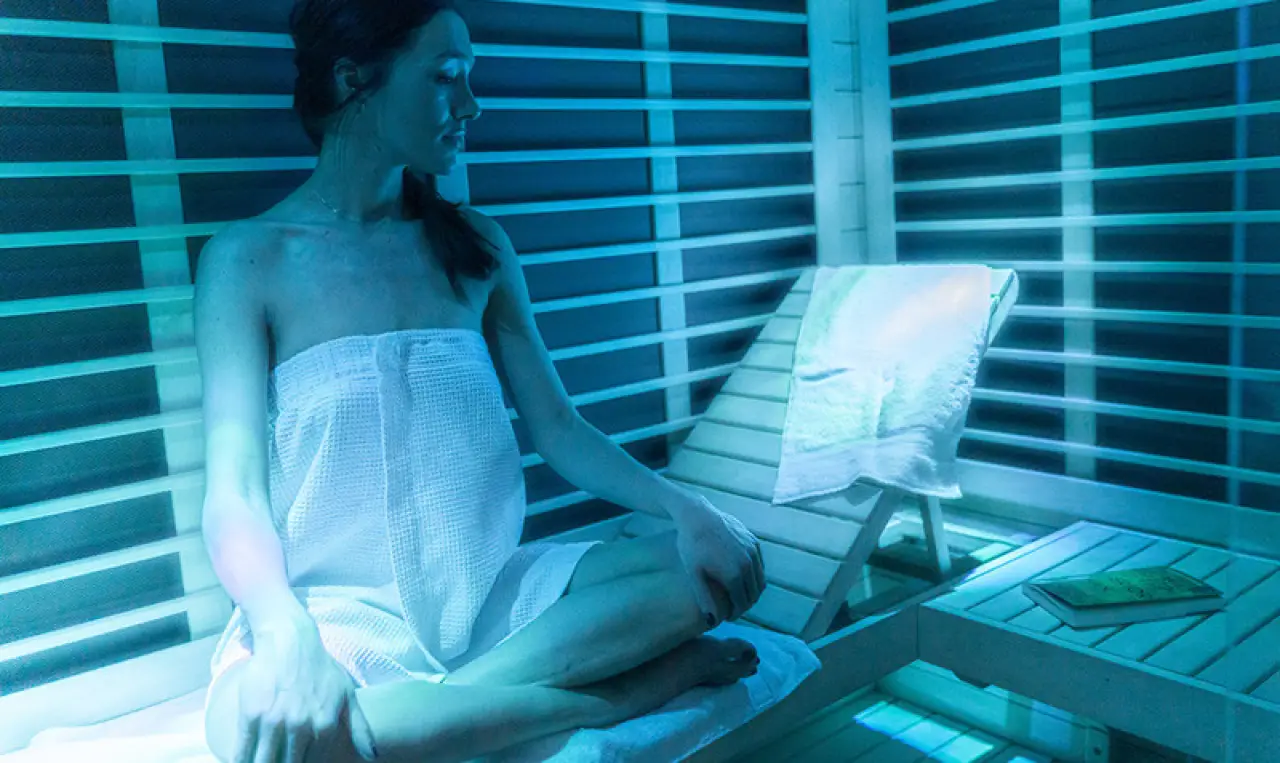 Woman in a sauna with a towel, sitting on a bench under blue lighting.