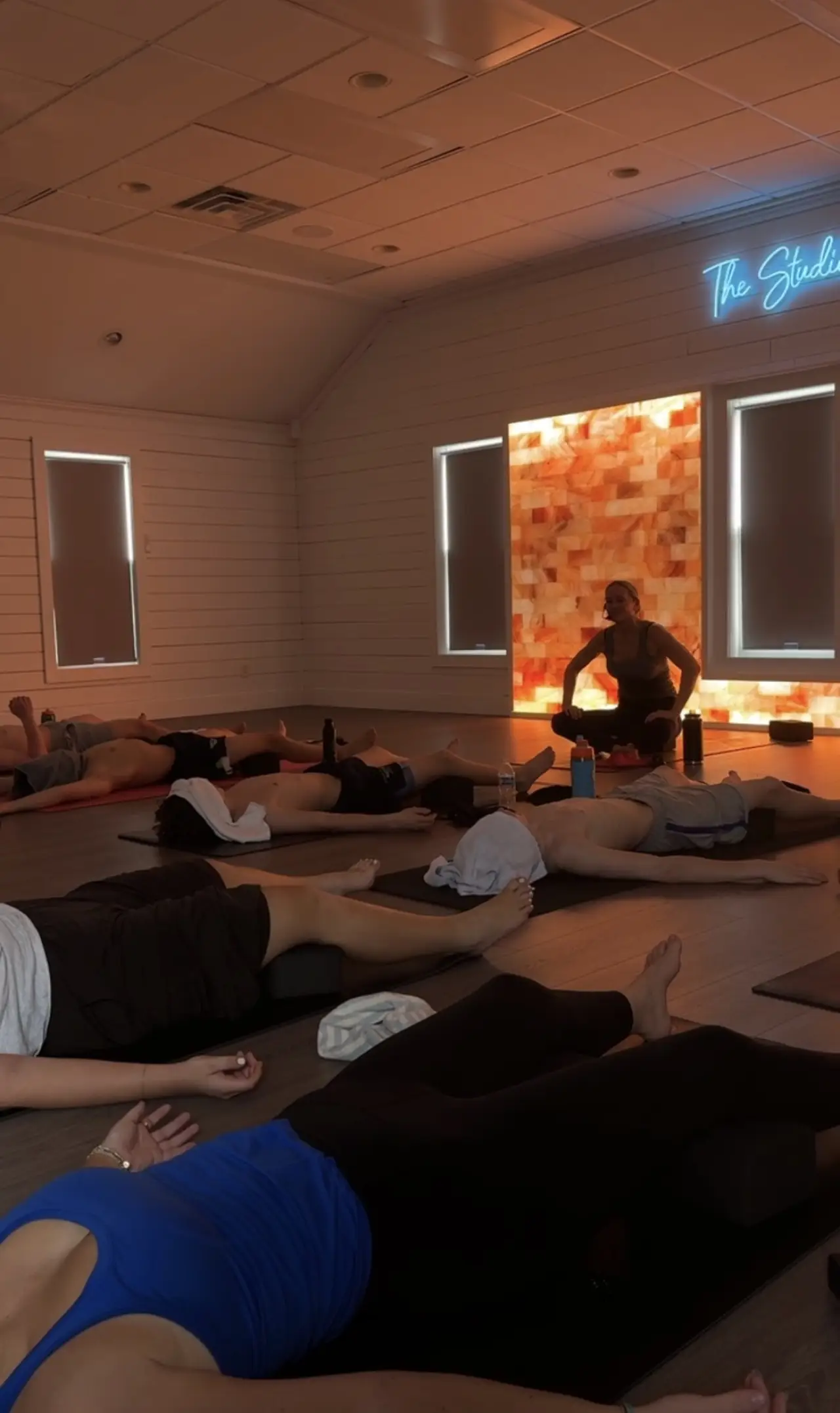 A yoga instructor assists a class of people lying on mats in a restful pose indoors.