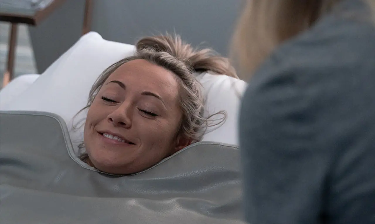 Person smiling while lying under a blanket, with someone standing nearby.