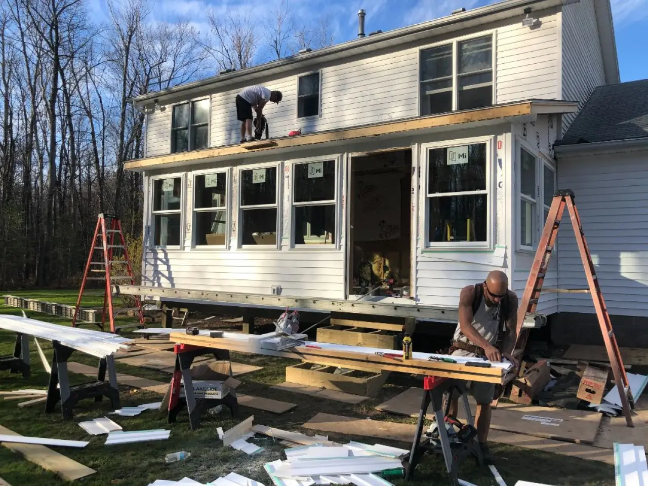 Two people work on house renovations; one cuts wood while another is on a ladder by windows.