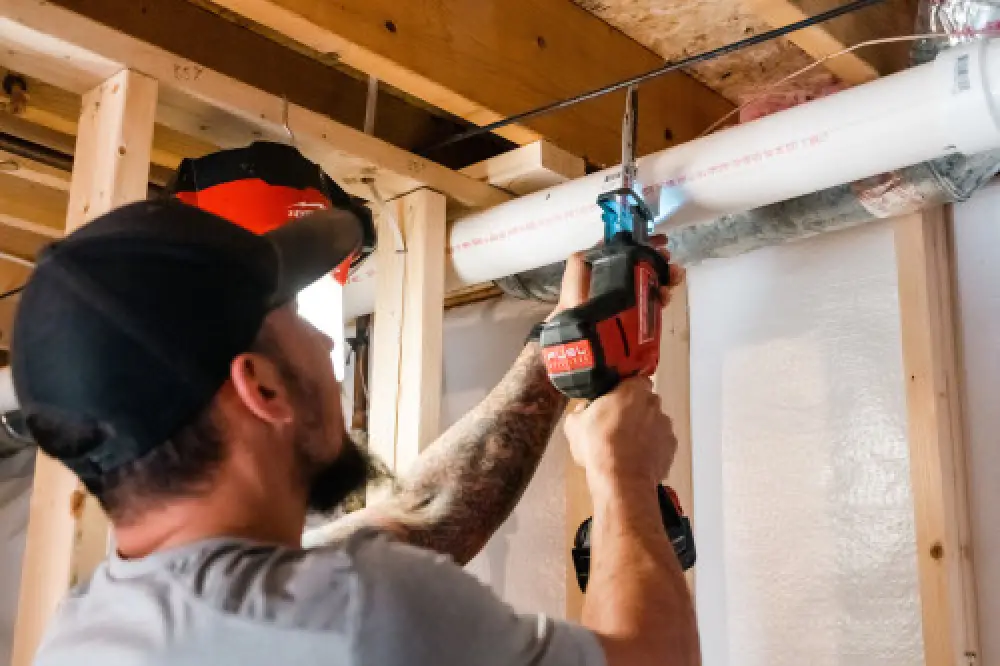 Man using a reciprocating saw to cut a white PVC pipe in a basement under construction.