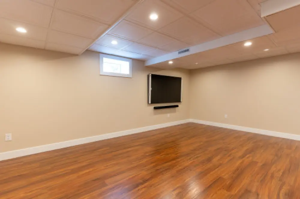 Empty basement room with wood flooring, beige walls, recessed lights, and a wall-mounted TV.