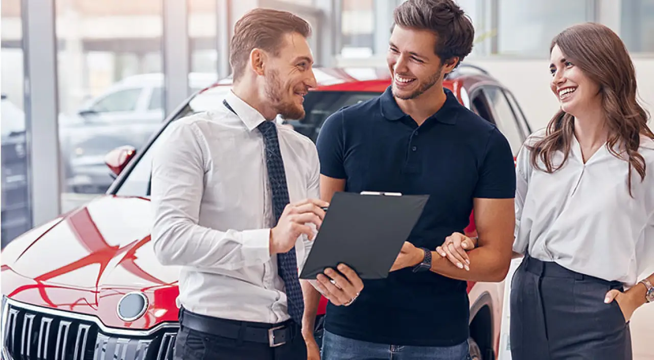 Three people smiling and talking in front of a red car at a dealership.