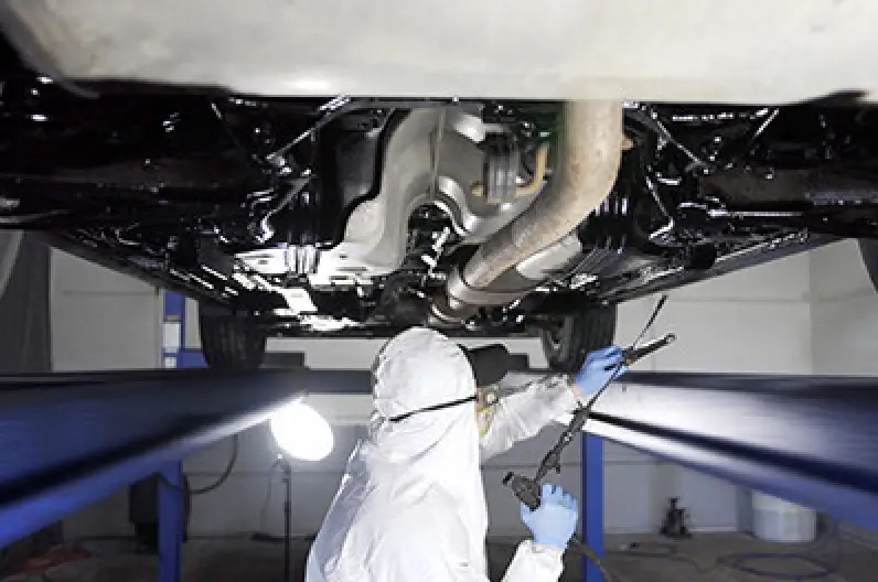 Person in protective gear sprays undercarriage of a car elevated on a hydraulic lift.