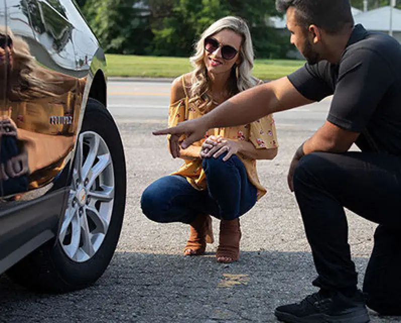 A man and woman kneel by a car, examining its tire and side in a parking lot.
