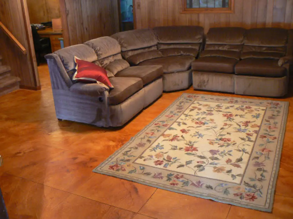 Brown sectional sofa with a red pillow and a floral rug on a wooden floor in a paneled room.
