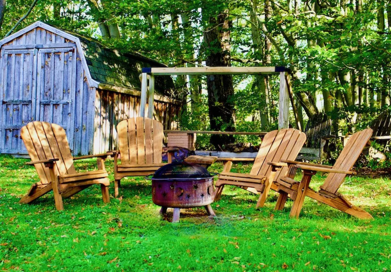 wooden Adirondack chairs around a fire pit in a lush garden with a shed and swing set in view