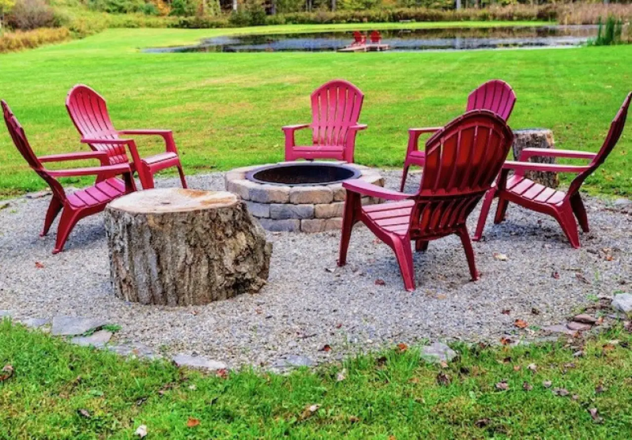 six red chairs are arranged around a fire pit on gravel, with a grassy field and pond nearby