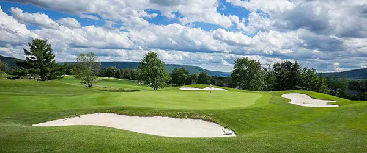 a golf course with sand bunkers, green grass, trees, and hills under a partly cloudy sky