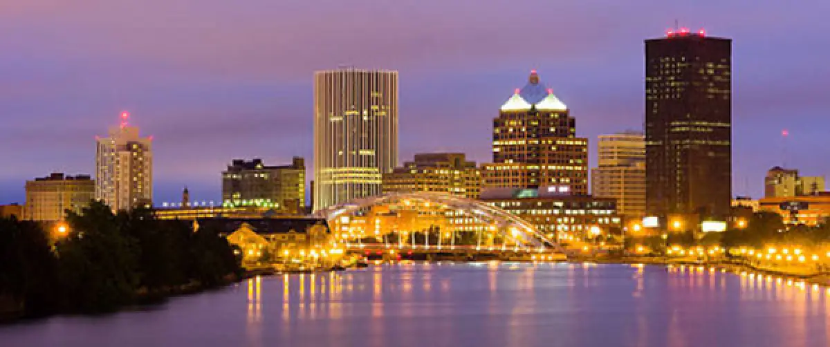 city skyline with lit buildings and a bridge reflected in a river at dusk