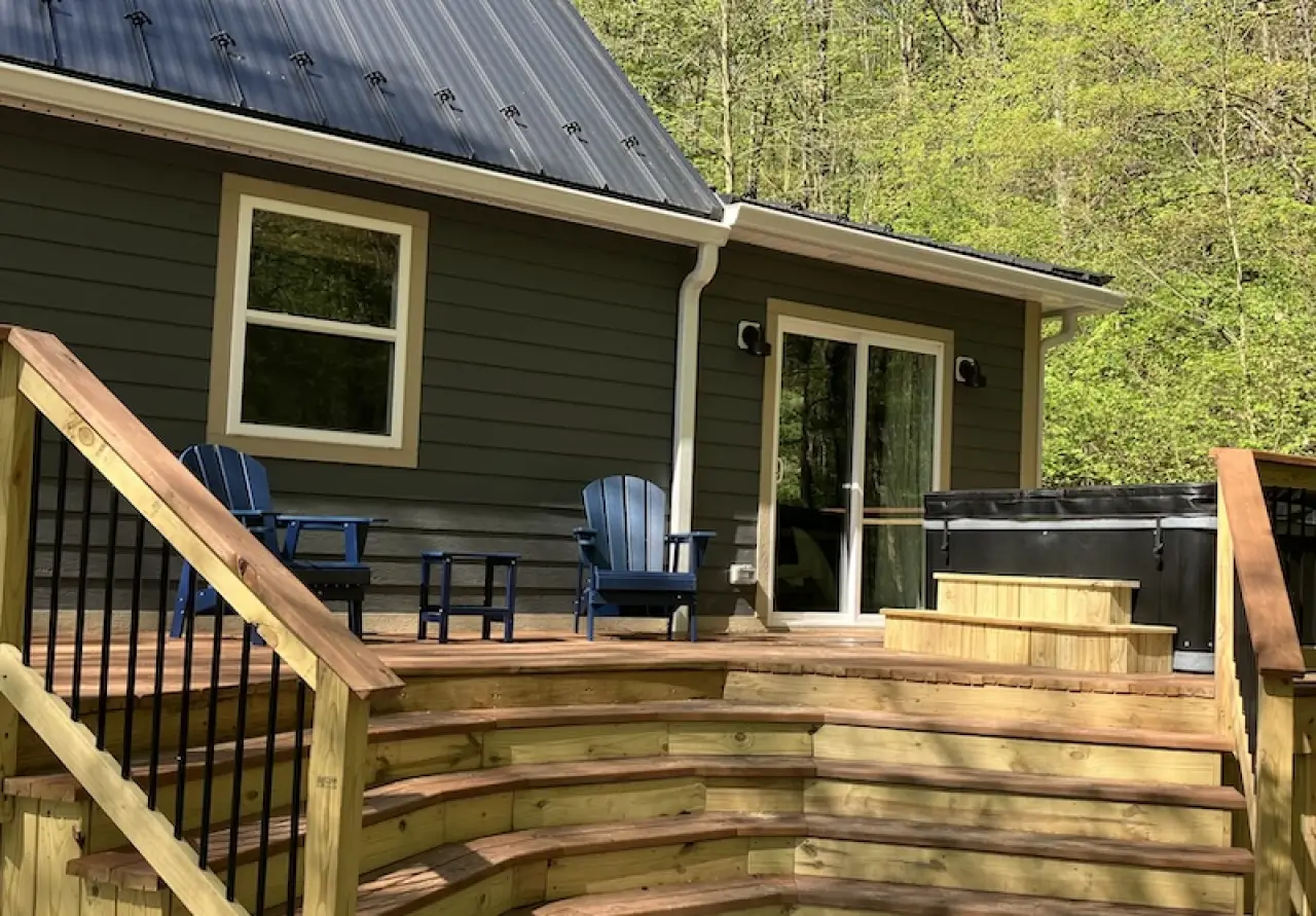 wooden deck with Adirondack chairs, hot tub, and a green house with forest backdrop
