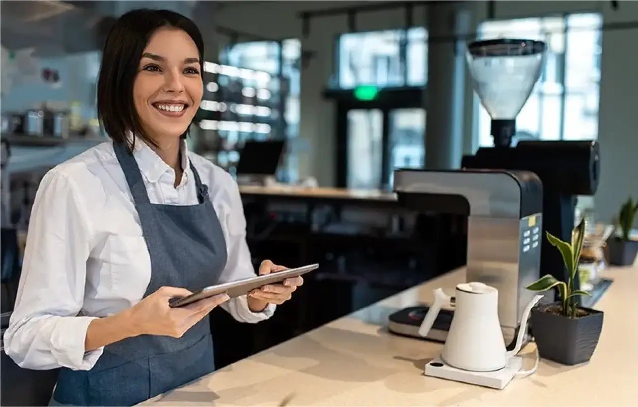 woman smiling working at a coffee shop holding an ipad and wearing an apron