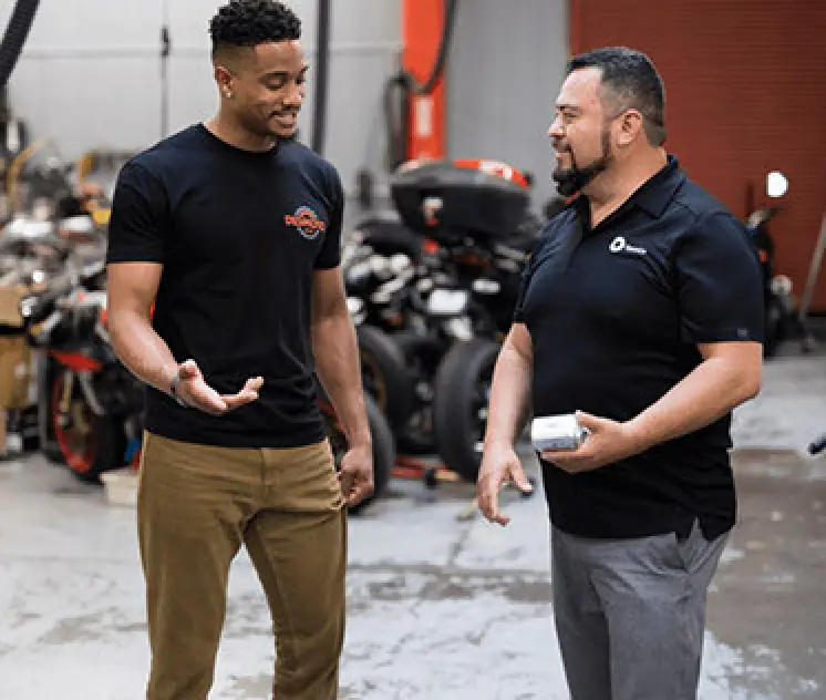 two men speak to each other outside with various small motor vehicles in background