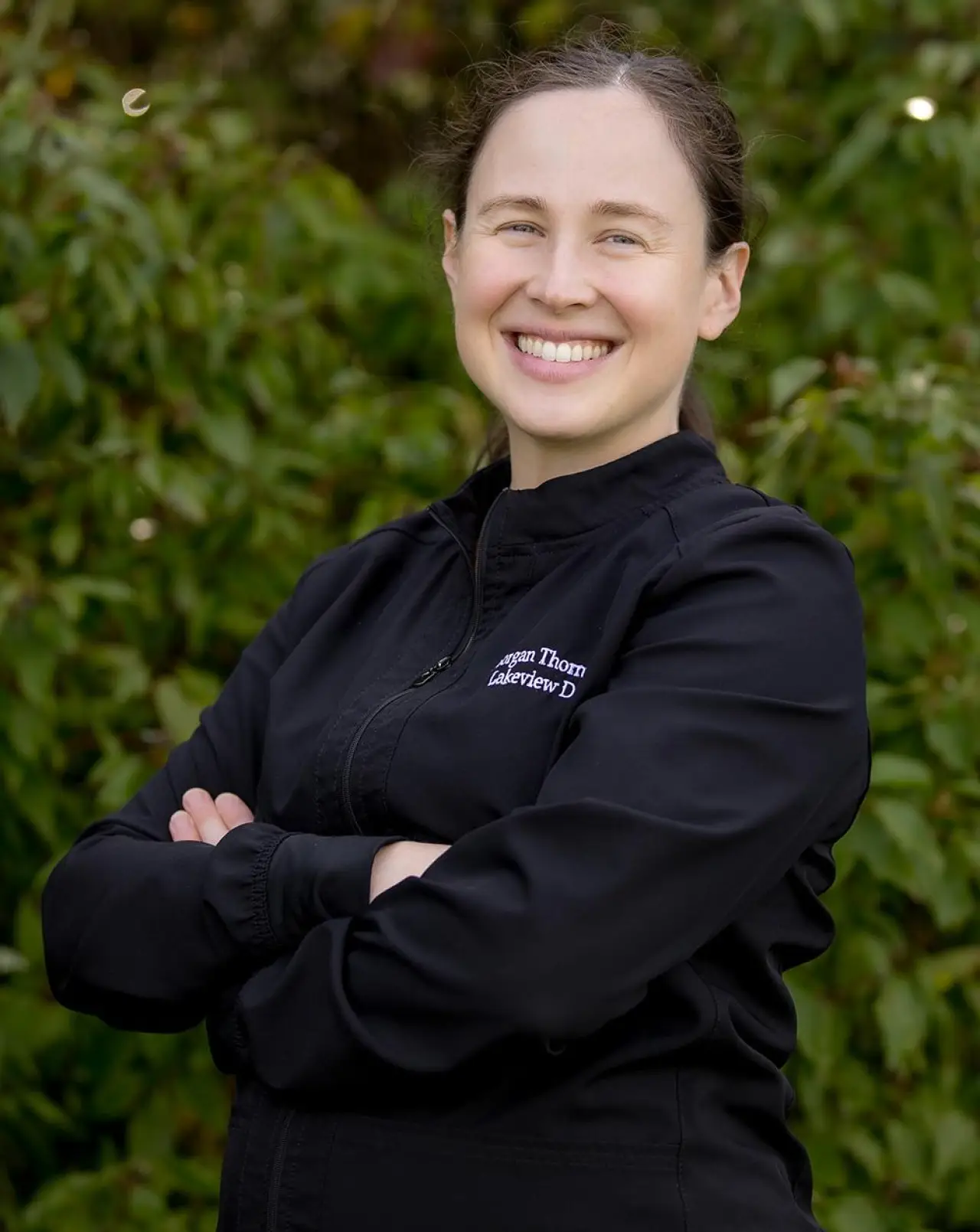 A smiling woman with arms crossed standing in front of green foliage.